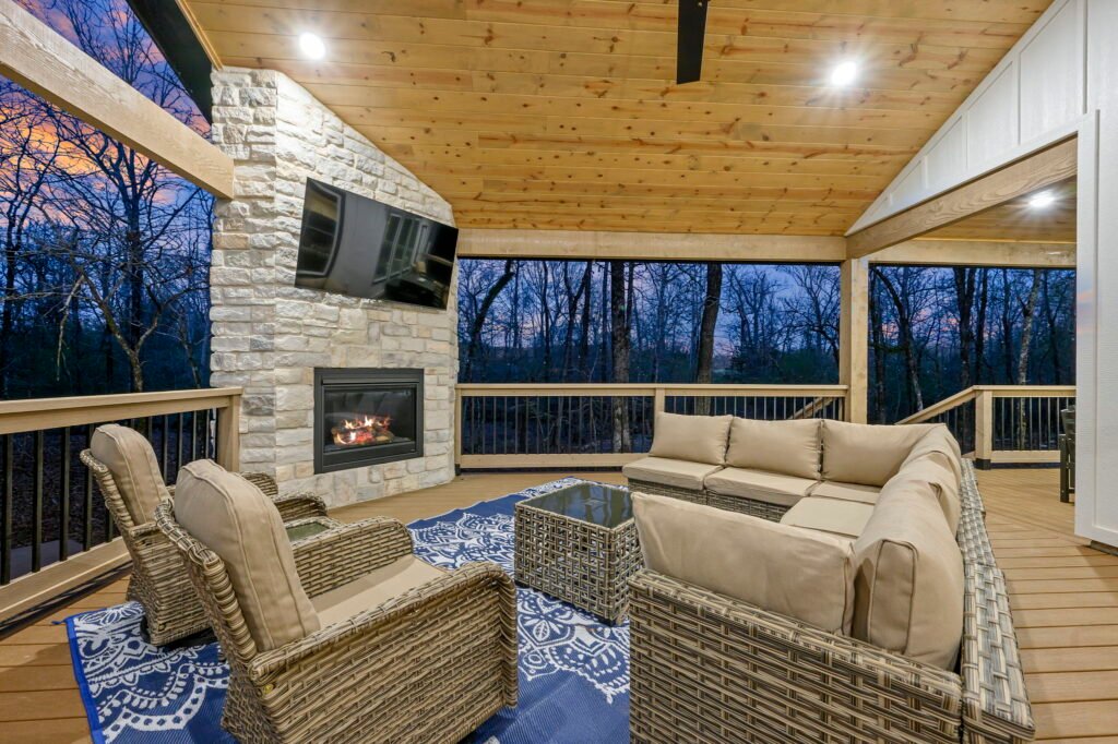 Covered outdoor patio with seating, fireplace, and TV overlooking forest at dusk