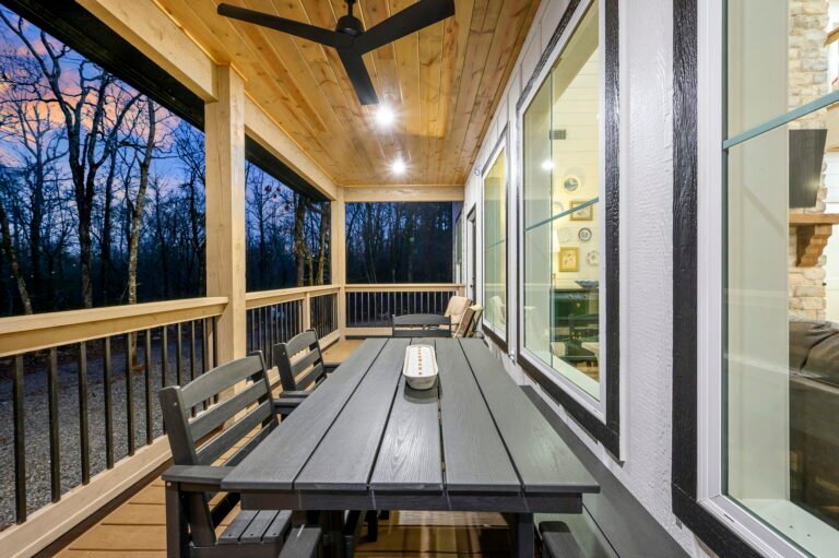Covered porch with outdoor dining table, ceiling fan, and forest view at dusk